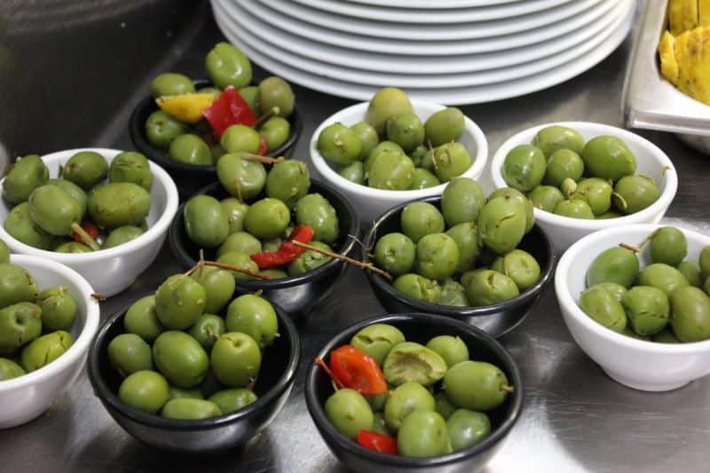 Tray of tapas full of small bowls of green olives, ready for serving, along with stack of plates in background