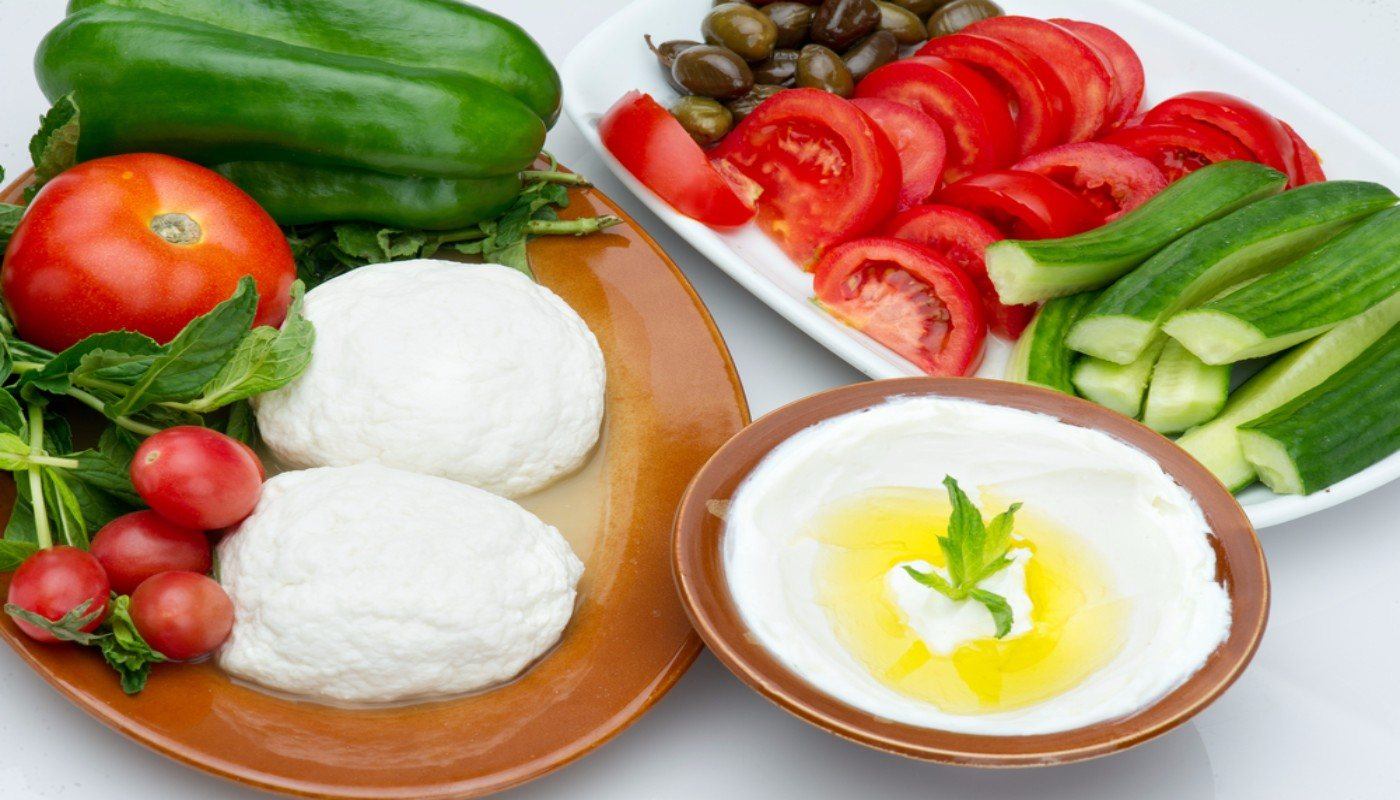 Bowl of Labneh surrounded by a platter and cutting board with cucumbers, herbs, radishes, olives and tomatoes.