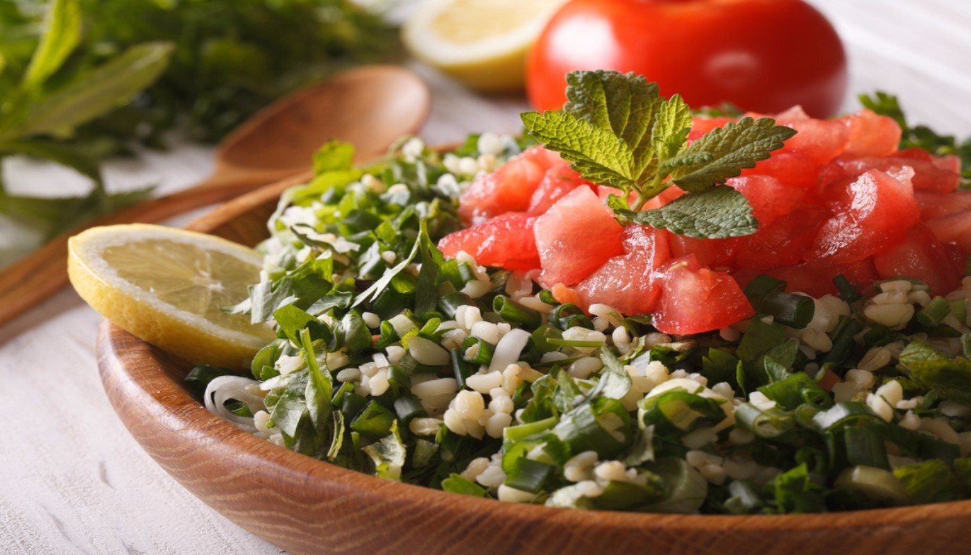 Bowl of tabouleh topped with tomatoes and a sprig of mint and garnished with a slice of lemon. Tomato in background.