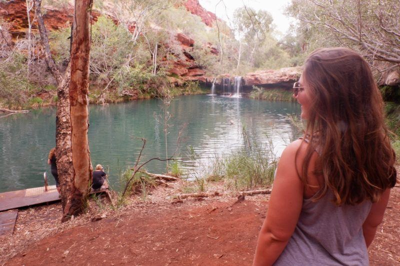 Woman standing in front of a lake with a small waterfall in the background.