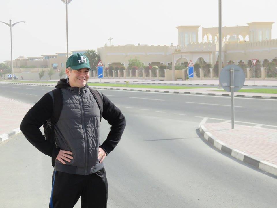 Ryan Biddulph stands along a road. Large building in the background.