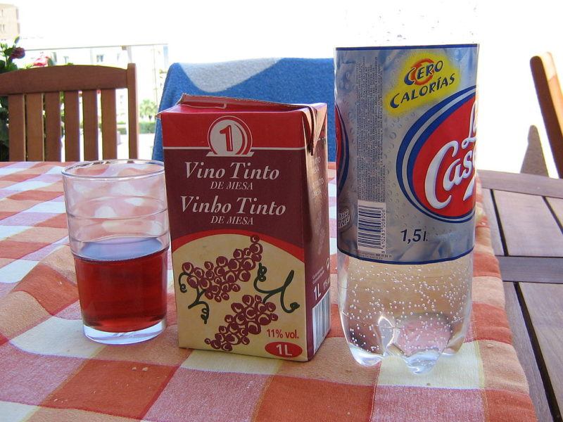 Small juice glass, box of Spanish red table wine, and bottle of La Casera, on a picnic table with red tablecloth.