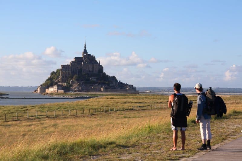 Two backpackers in foreground looking at Mont Saiint-Michel. Grass and water between them.