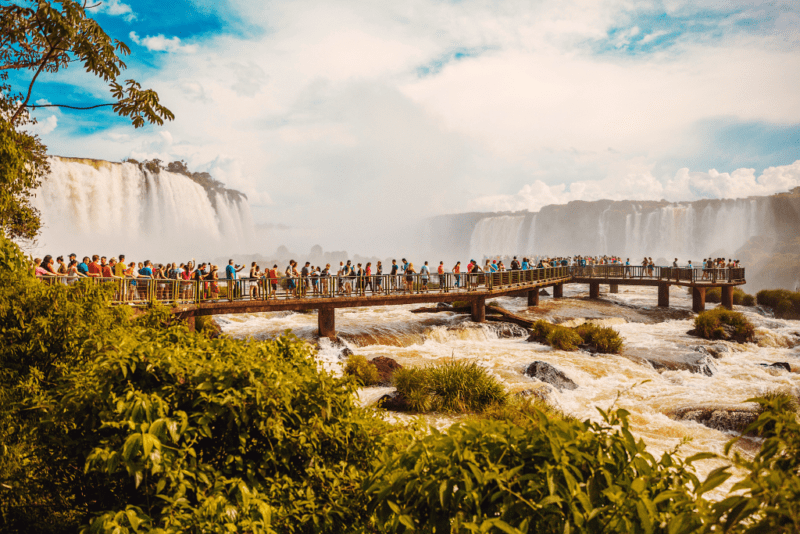 crowds of people stand on walkway over the water coming from Iguazu Falls in the background