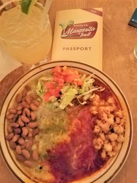 Plate of enchiladas topped with green and red chili. Sides are corn, beans and chopped lettuce & tomato. Margarita Trail Passport and margarita in background.