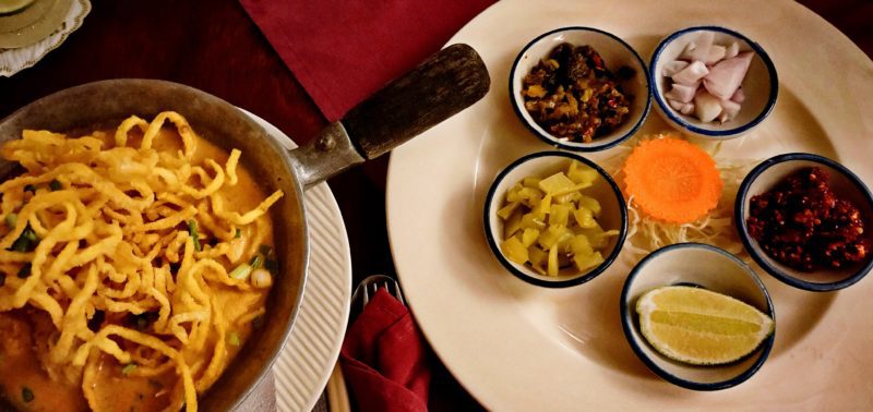 Bowl of Khao Soi Thai food with plate holding small bowls of soup accompaniments