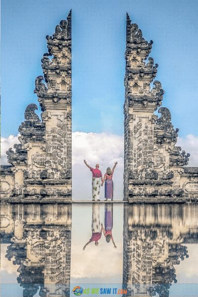 A couple stand at the candi bentar split gate at Pura Lempuyang temple in Bali, with their outer hands raised. Image reflected at ground level.