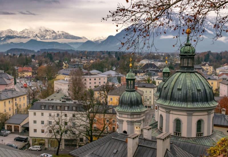 Rooftops of Salzburg taken from a hilltop. Tree in foreground, snow-capped mountains in background.