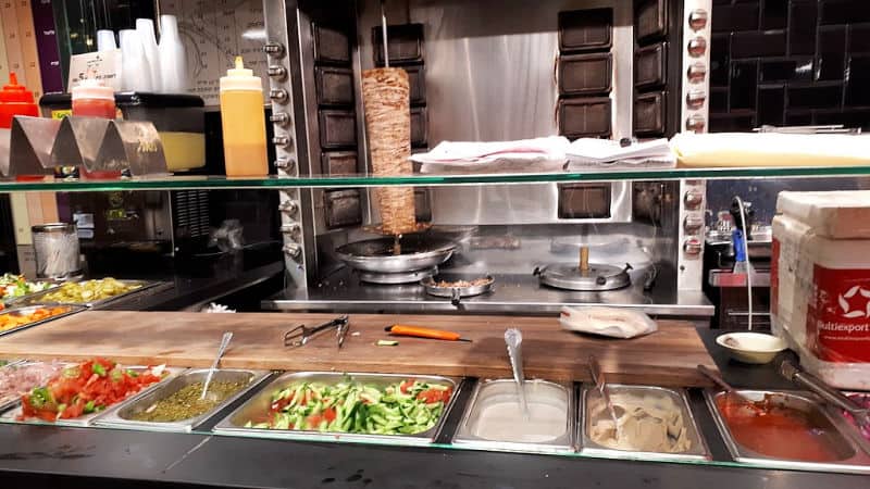 Healthy Arabic foods restaurant counter. Shawarma meat on the spit in background. Foreground: prep station with assorted accompaniments