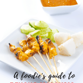 Chicken satay skewers on a plate with cucumber and rice cakes. Bowl of peanut sauce nearby. Text overlay says "A foodie's guide to Penang Malaysia"