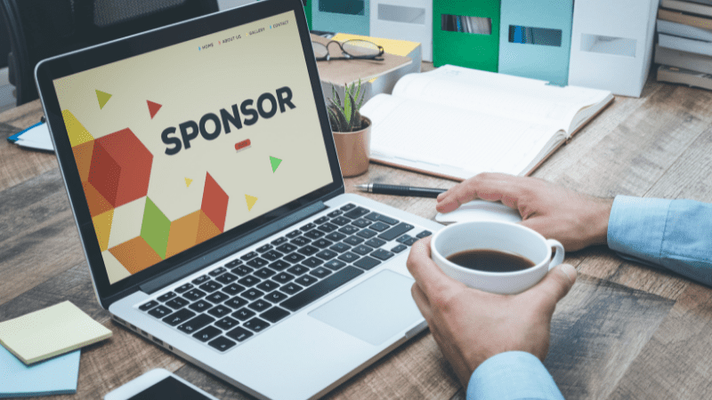 Computer with the word sponsor on the screen. Hand holding coffee cup. Symbolizing sponsored content and sponsored posts on our travel blog