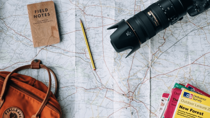 Learning how to start a travel vlog begins with having the right travel blogger gear. In this image: map, pencil, notebook, camera, and bag.