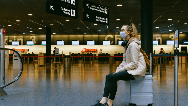 Typical post-pandemic travel trend: woman sitting on a suitcase in an airport, and wearing a mask