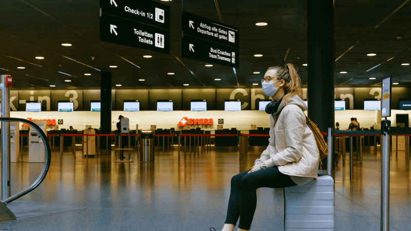 woman sitting on a suitcase in an airport, wearing a mask