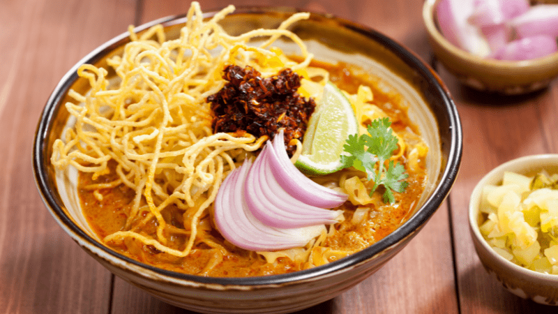 khao soi thai noodle soup topped with onion slices, chilies, a lime wedge, and a sprig of cilantro