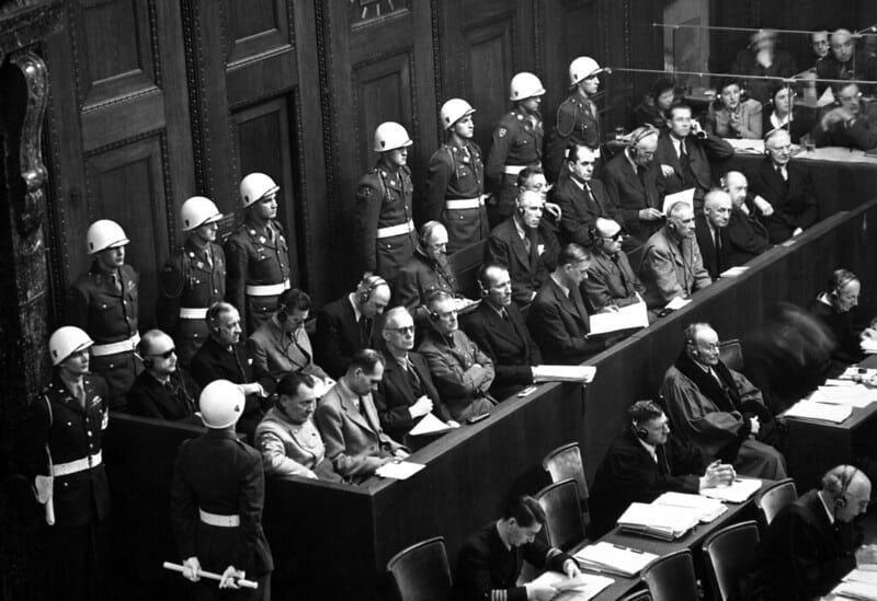Black-and-white vintage photo of men sitting in chairs at the Nuremberg Trials. Military Police line the room.