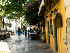 Buildings lining an ancient cobbled road in Corfu. Tables and people on the pedestrian pathway, with bougainvillea and ivy overhead