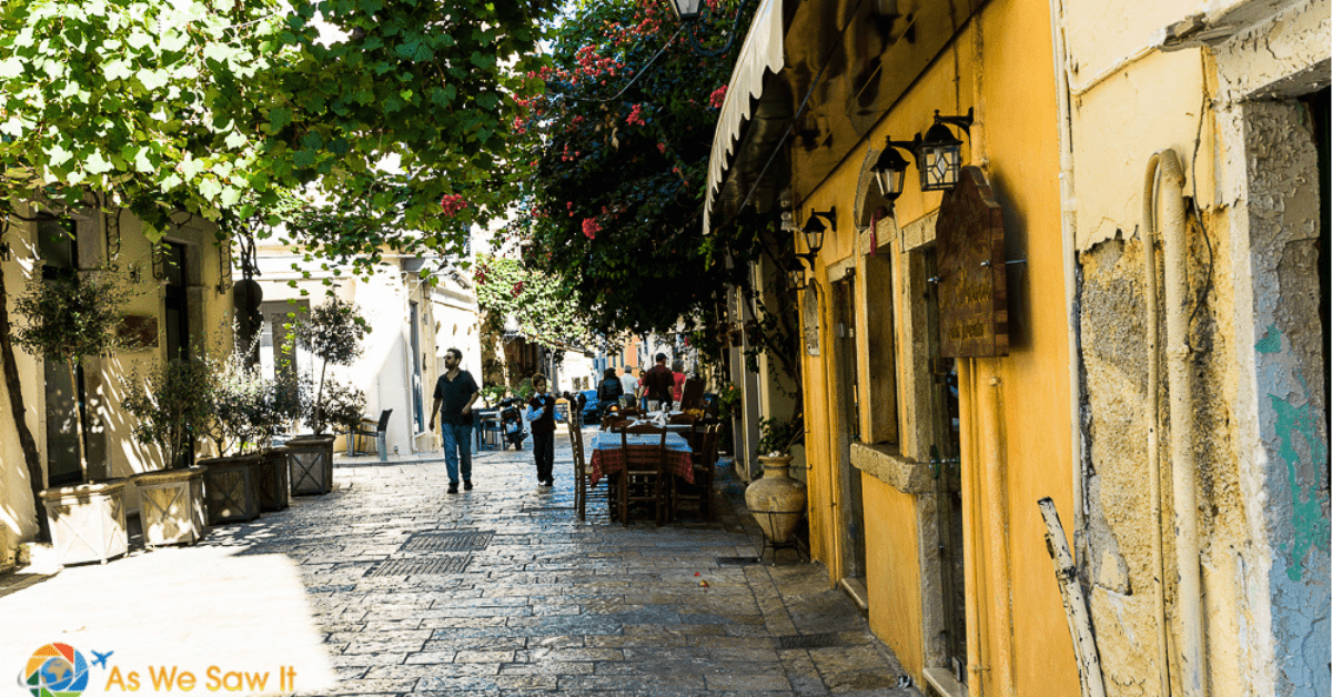 Buildings lining an ancient cobbled road in Corfu. Tables and people on the pedestrian pathway, with bougainvillea and ivy overhead