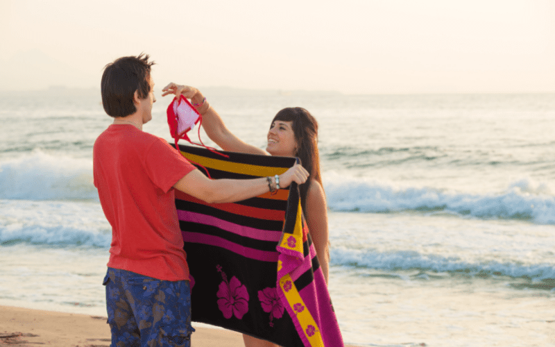man holds a towel for a woman changing clothes at the beach