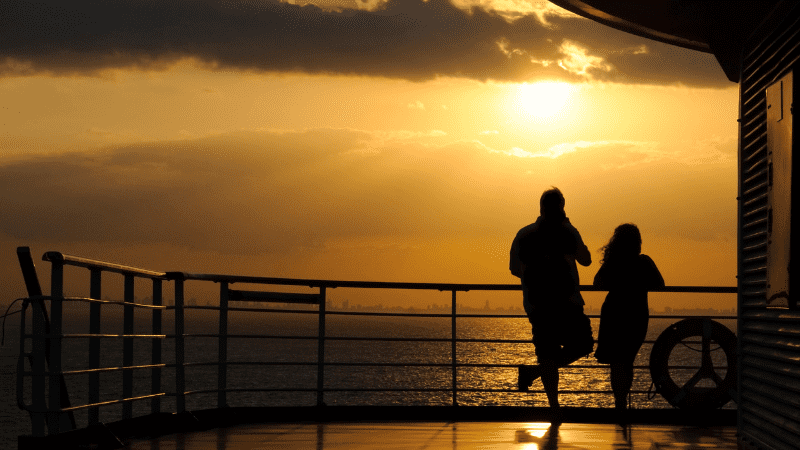 man and woman standing at the rail of a ship, with ocean and sun in background