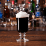 Mug of Irish coffee with shelves full of colorful bottles in background.