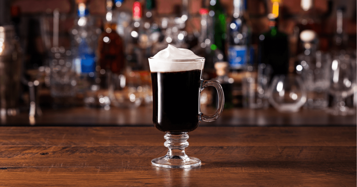 Mug of Irish coffee with shelves full of colorful bottles in background.