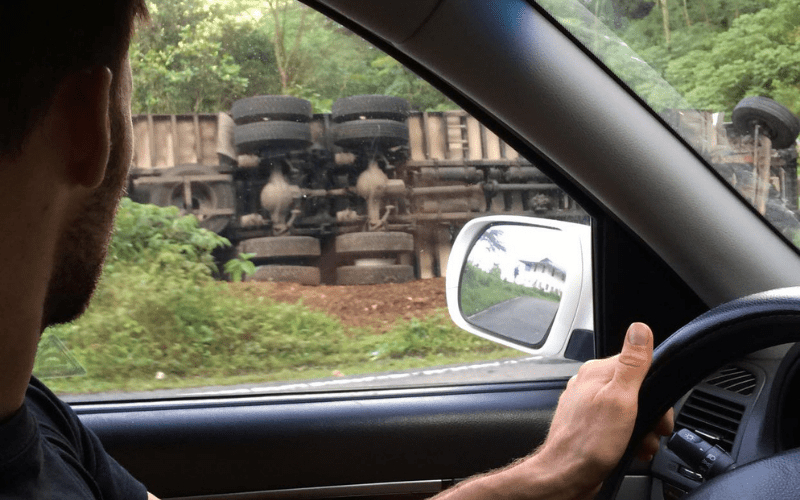 Man in a car looking at a truck that flipped over on the side of a road.