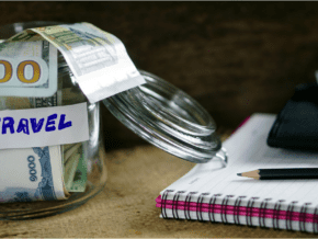 Jar full of money with a travel label on it. Notebook with pen. How to save money on travel