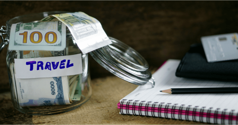 Jar full of money with a travel label on it. Notebook with pen. How to save money on travel