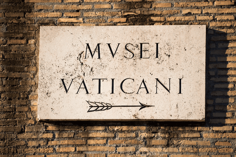 Sign with an arrow pointing to the right below the Latin words MVSEI VATICANI, showing location Vatican Museums. 