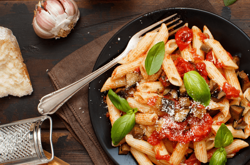 a plate of penne pasta alla Norma, with a fork, garlic, and parmesan cheese with grater.
