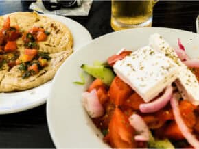 Greek salad topped with a hunk of feta cheese. Plate of pita bread on the side