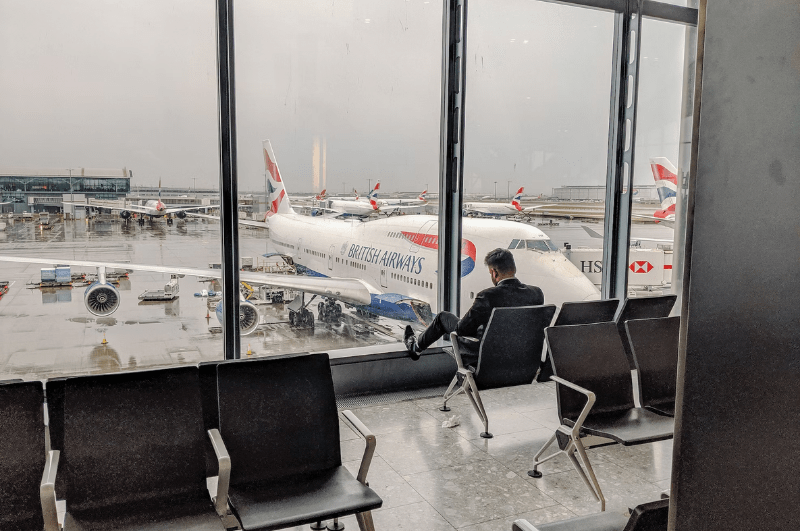 Man during a layover in London, sitting in a chair in an airport with British Airways plane outside the window.