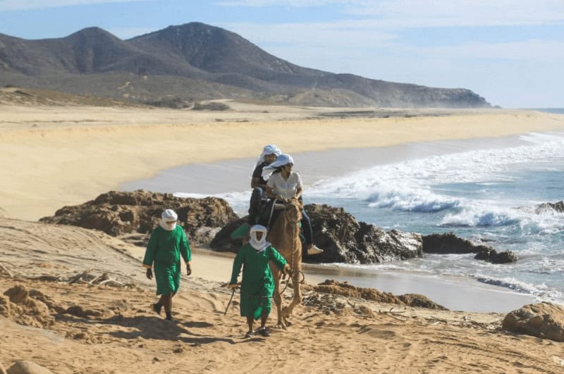 Two people leading a couple on a camel on the beach.