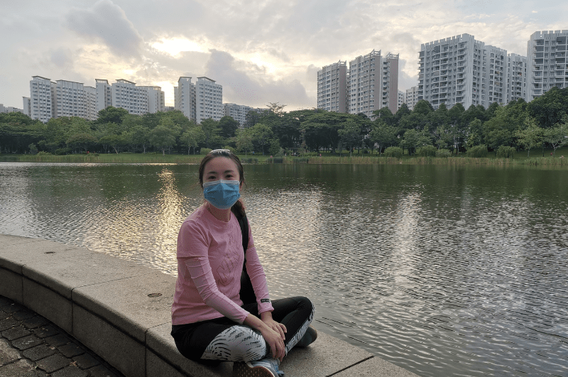 Woman wearing a mask, sitting on a wall in Punggol Park. Water and buildings in background