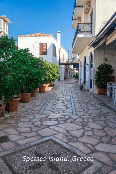 Picturesque pedestrian walkway lined with potted plants and homes. Text overlay says "Spetses Island Greece.