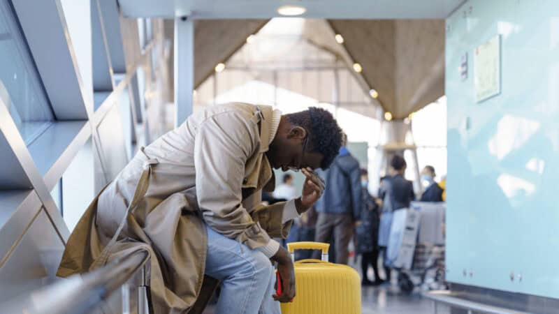 A man sitting on a windowsill at the airport, with his head in his hands. Yellow suitcase next to him.