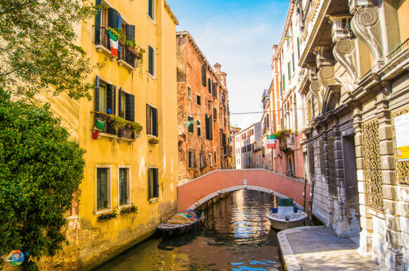 A local canal in Venice, spanned by a bridge and framed by flower filled balconies.