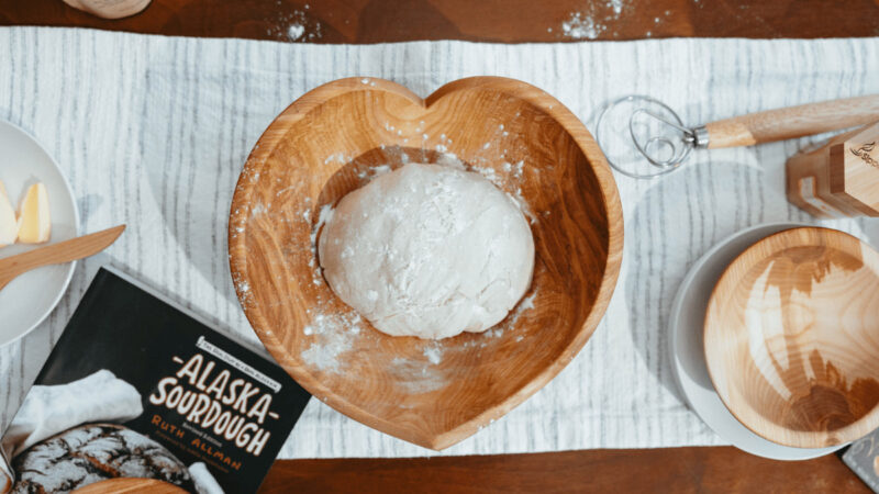 Heart-shaped birch bowl on a table, holding bread dough. Also on the table: an additional bowl, Alaska Sourdough cookbook, and assorted kitchen tools.