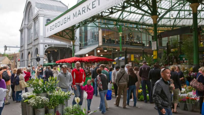 Crowds browsing stalls under the green metal canopy of Borough Market in London, with fresh flower buckets in the foreground and red umbrellas covering vendor tables.