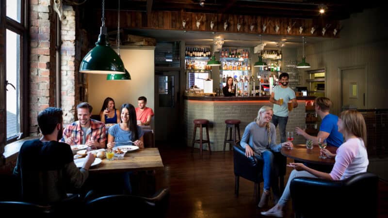 Warmly lit British pub with exposed brick walls and wooden beams, featuring patrons dining and chatting at tables while a bartender serves drinks behind a stocked bar.