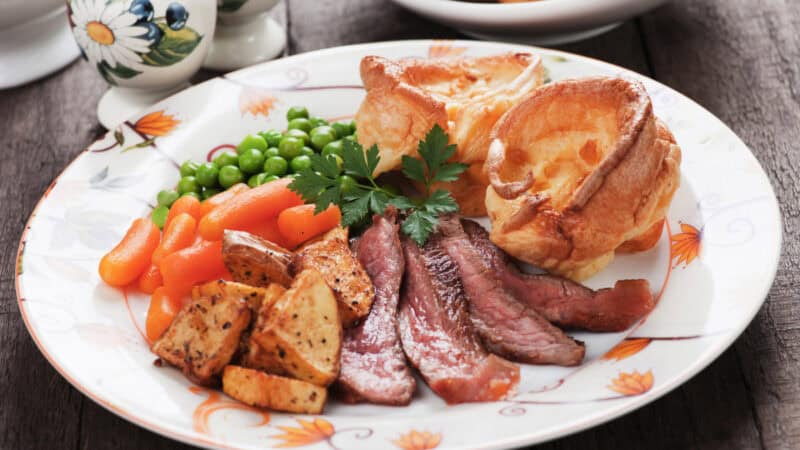 Decorative plate with roast beef slices, herb-roasted potatoes, baby carrots, green peas, Yorkshire puddings, and parsley garnish on a wooden table.