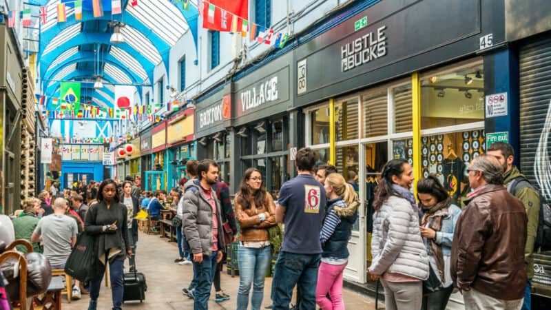 Busy indoor market arcade in Brixton, London, with blue arched ceiling, international flags, and diverse crowd walking past shops and eateries like ‘Hustle Bucks’ and ‘Village.’