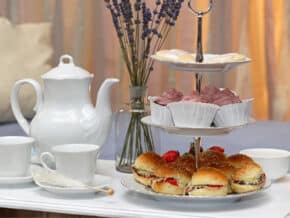 White porcelain teapot and teacups with a two-tiered stand holding frosted cookies and cupcakes, plus cream-filled strawberry buns, set on a white table with lavender vase and soft blue couch in the background.