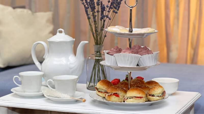 White porcelain teapot and teacups with a two-tiered stand holding frosted cookies and cupcakes, plus cream-filled strawberry buns, set on a white table with lavender vase and soft blue couch in the background.