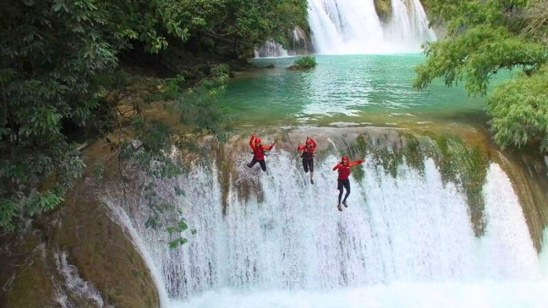 Adventurers leap off a rocky ledge into a turquoise waterfall pool, framed by lush jungle and cascading tiers.