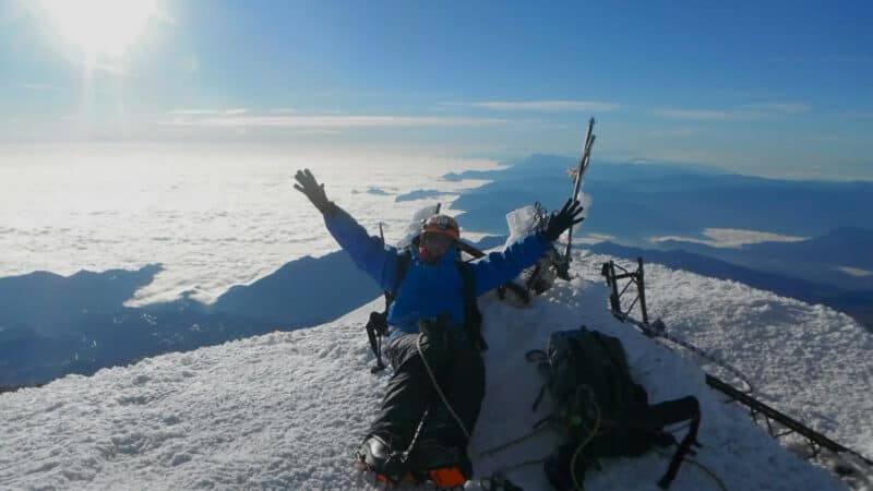 A climber in mountaineering gear raises their arms in triumph atop a snow-covered peak above the clouds.