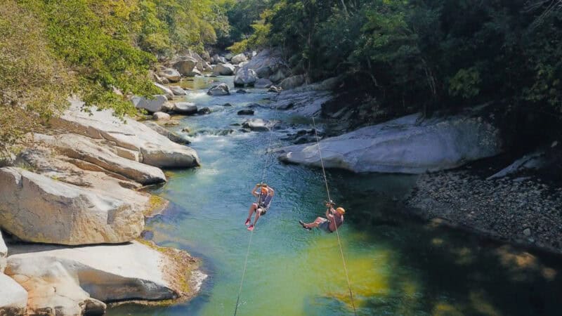 Two people zip-line over a green-hued river surrounded by boulders and dense forest. Location: Puerto Vallarta, one of the best adventure spots in Mexico