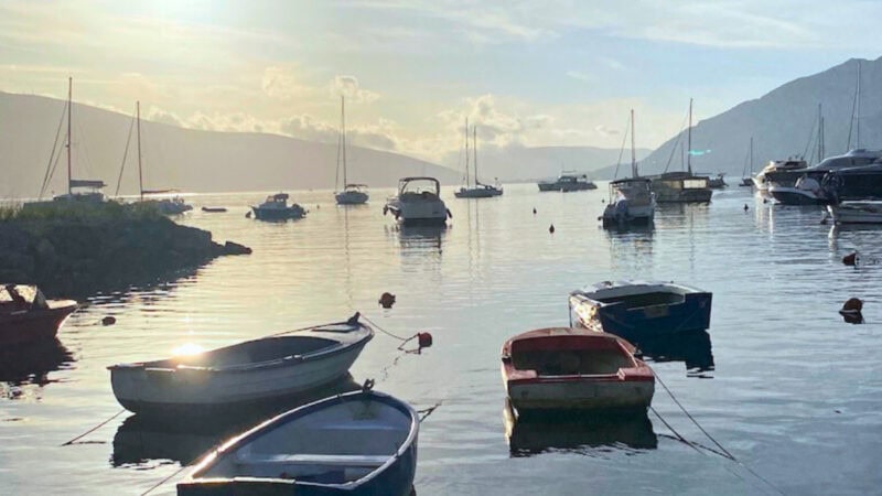 Serene sunset or sunrise view over the Bay of Kotor, with several small, simple fishing boats moored in the foreground and a collection of larger sailboats and yachts anchored further out, all against a backdrop of shadowed mountains.