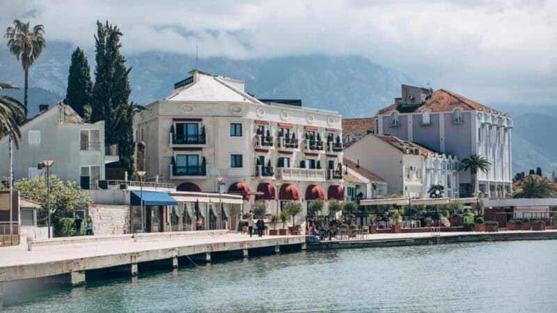 The luxurious white and terracotta buildings of Porto Montenegro in Tivat, with red awnings and outdoor seating, stretching along the calm, sunlit waterfront with a large mountain range behind them.